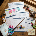 A collection of Great Wolf Lodge travel planning documents, a tablet displaying an instant download page, a child's swimsuit, a toy wand, five small family figurines, sunglasses, a bag of trail mix, a plush wolf, and markers.