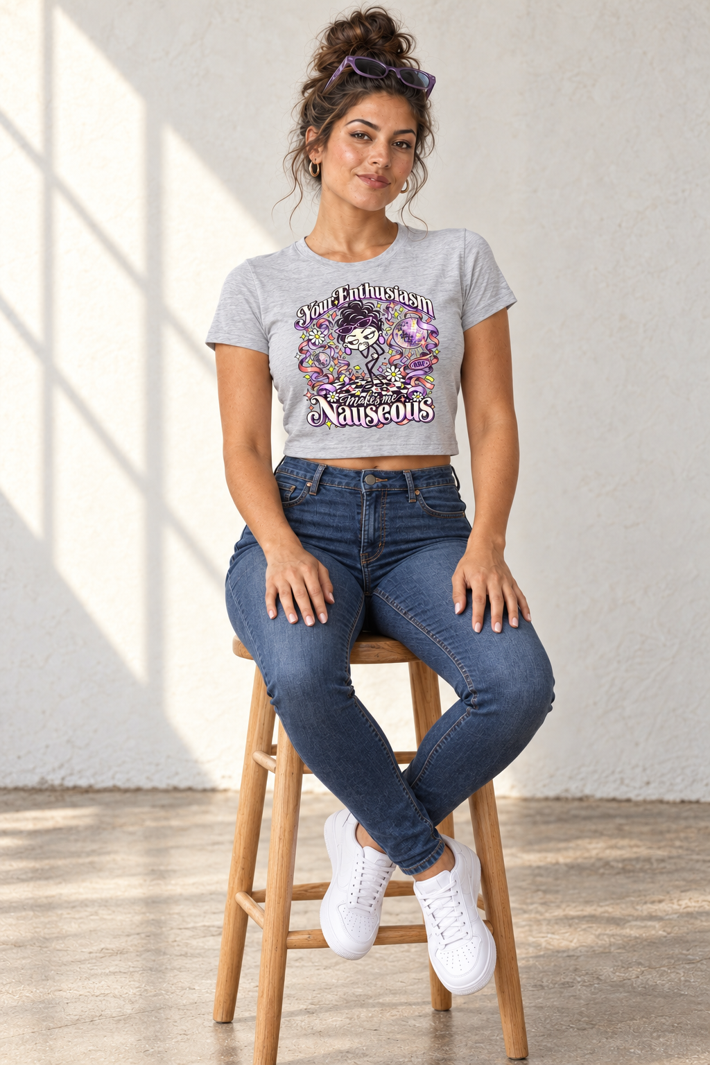Lifestyle mockup: Your Enthusiasm Makes Me Nauseous crop tee in Athletic Heather. Seated on a studio stool with the front design visible.