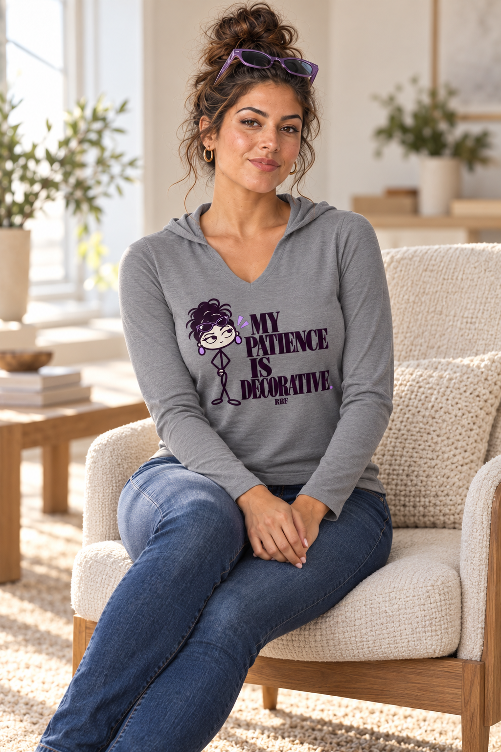 Lifestyle mockup: My Patience Is Decorative hooded long-sleeve top in Grey Frost. Seated in a sunlit lounge with the front design visible.