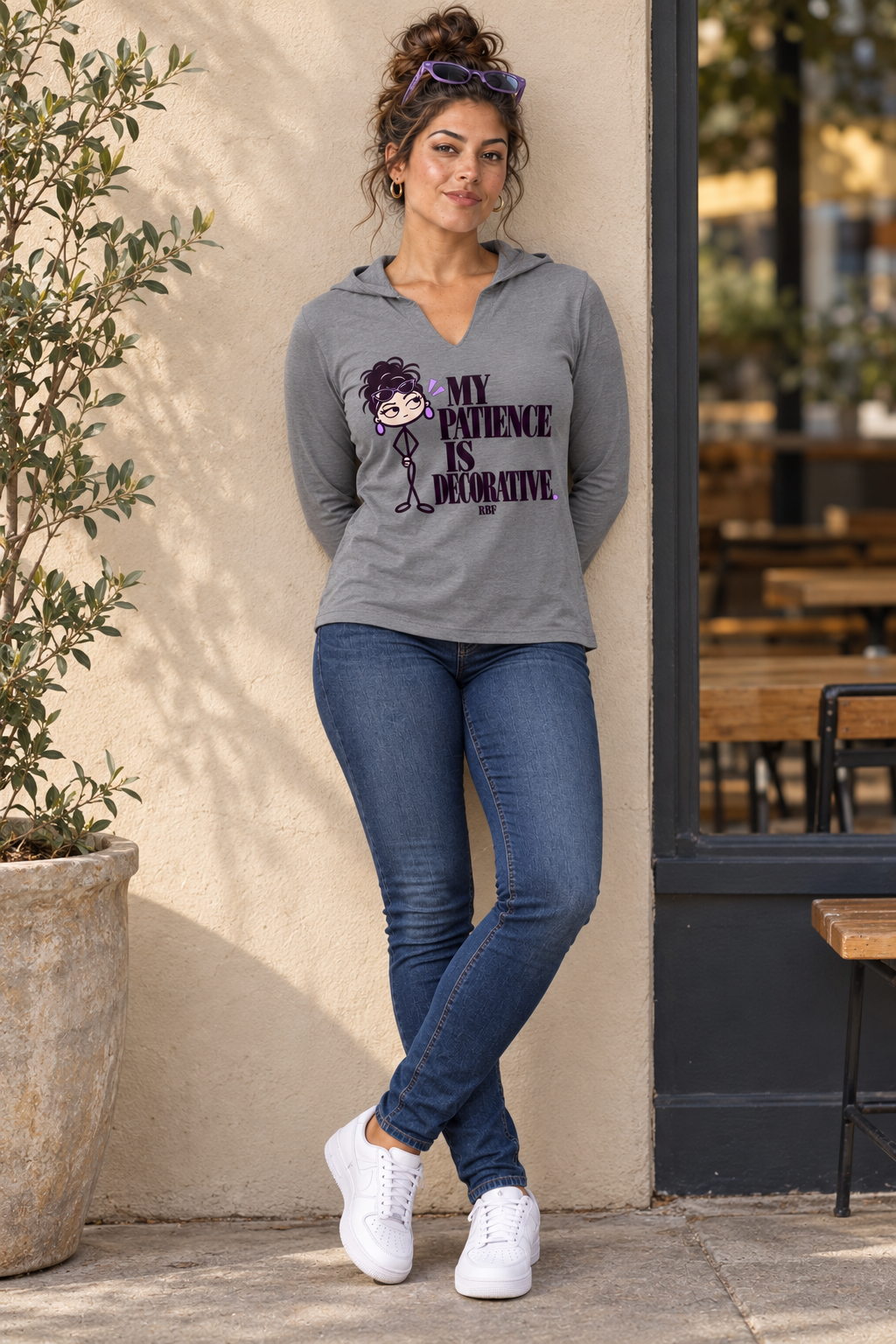 Lifestyle mockup: My Patience Is Decorative hooded long-sleeve top in Grey Frost. Leaning against a cafe wall with the front design visible.